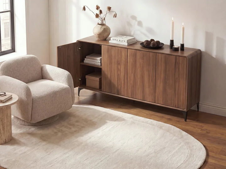 A mid-century modern wooden sideboard cabinet in a warm walnut finish, styled in a bright living room next to a beige armchair and a cream rug.