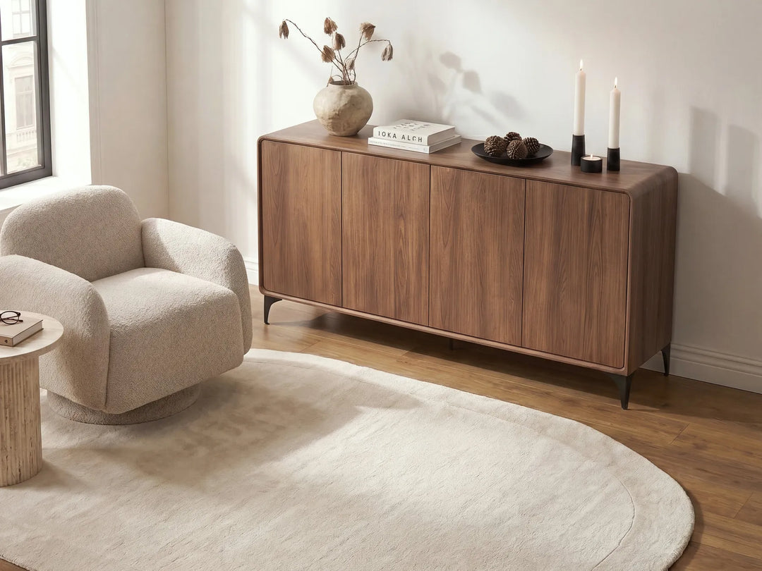 A mid-century modern wooden sideboard cabinet in a warm walnut finish, styled in a bright living room next to a beige armchair and a cream rug.