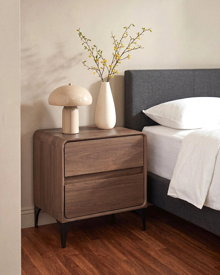 A modern two-drawer nightstand in a medium brown wood finish, styled next to a bed with a dark gray headboard and white bedding. The nightstand is decorated with a cream mushroom-shaped table lamp and a matching cream vase holding delicate yellow-brown branches.