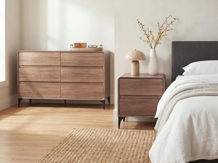 A modern bedroom scene featuring a six-drawer wooden dresser and matching nightstand in a warm medium brown finish, styled with a beige mushroom lamp, white vase with branches, and a white bed.