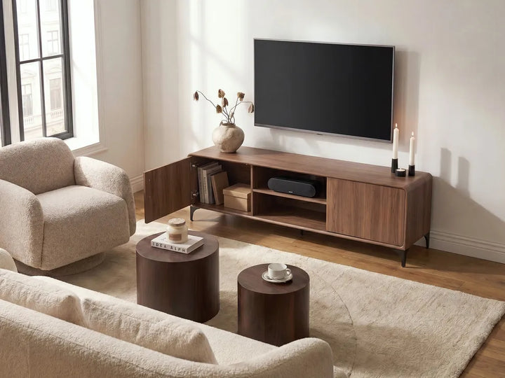 A modern wooden TV stand in a warm medium brown finish, styled in a bright living room with a beige armchair, a sofa, two dark wood coffee tables, and a wall-mounted flat-screen TV.