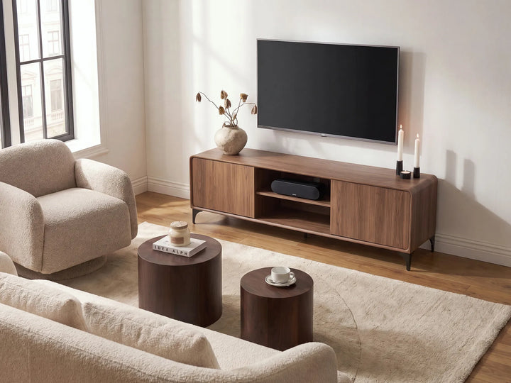 A modern living room scene featuring a dark wood TV stand console beneath a wall-mounted flat-screen TV, styled with a beige armchair, round wooden side tables, a light rug, and neutral decor.
