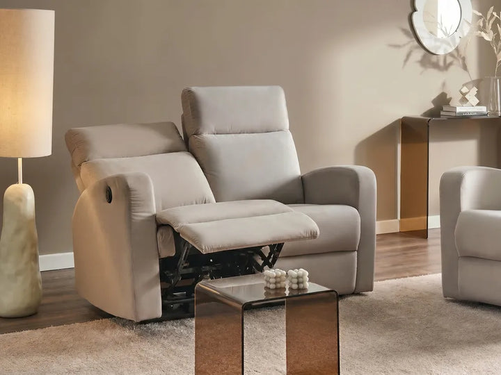 A light beige recliner chair with the footrest extended, shown in a styled living room setting with a floor lamp, side table, and another matching chair partially visible.