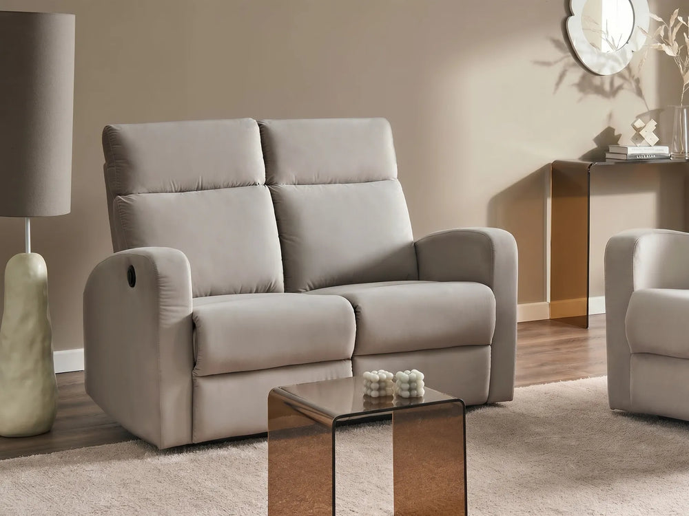 A light beige upholstered reclining loveseat sofa shown in a styled living room setting with a matching side table, lamp, and another partial chair visible.