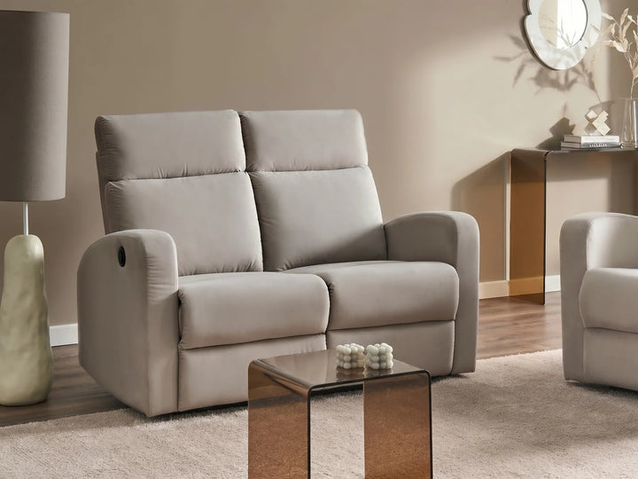 A light beige upholstered reclining loveseat sofa shown in a styled living room setting with a matching side table, lamp, and another partial chair visible.