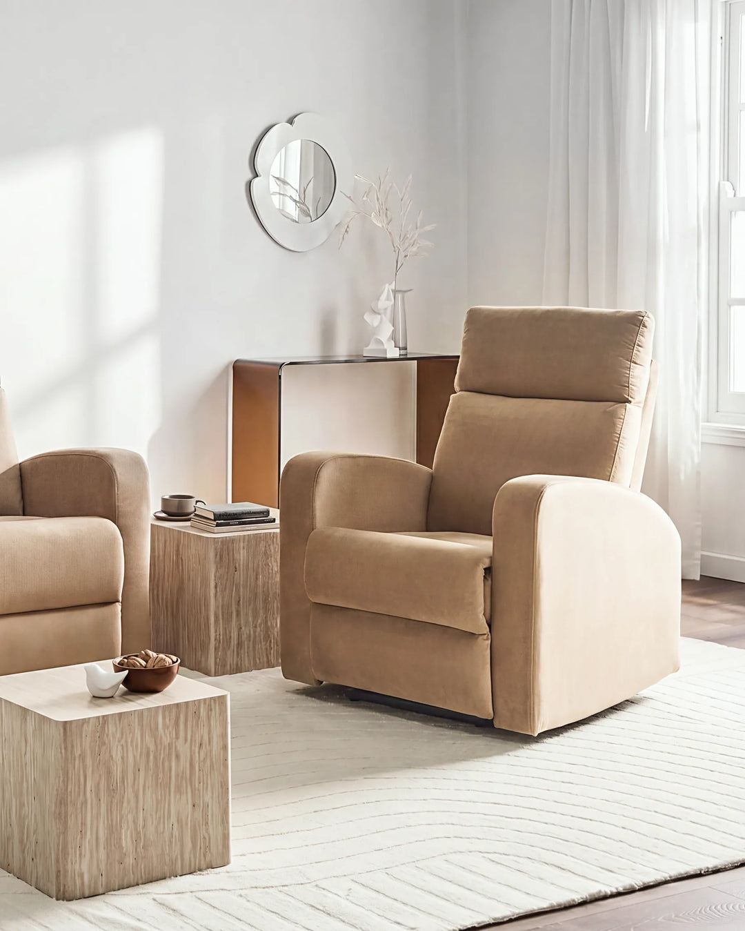 A beige upholstered recliner chair in a bright, modern living room setting with a matching armchair, wooden side tables, and a light rug.