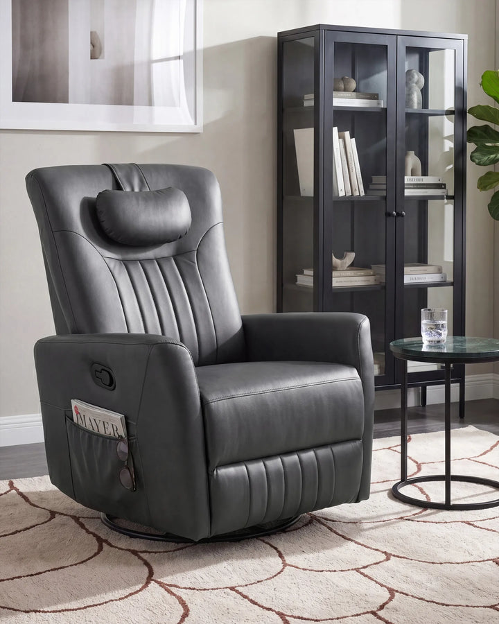 A dark gray upholstered recliner chair with a headrest and side magazine pocket, shown in a staged living room setting with a glass cabinet, side table, rug, and wall art.