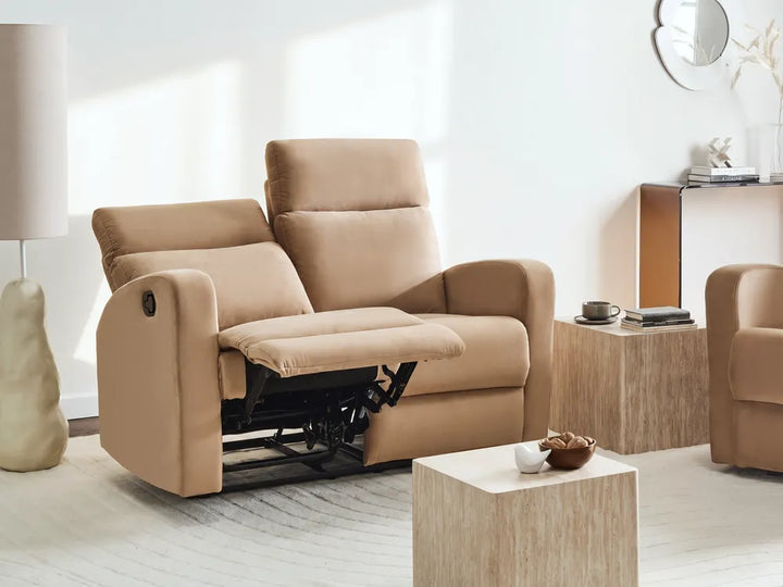A beige upholstered recliner loveseat with the footrest extended, shown in a bright modern living room setting with a side table, lamp, and another matching chair partially visible.