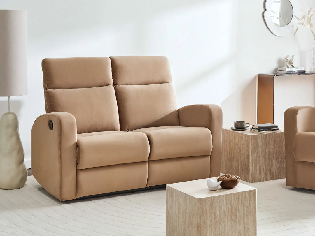 A beige upholstered reclining loveseat sofa shown in a bright, modern living room setting with a matching armchair, wooden side table, and decorative lamp.