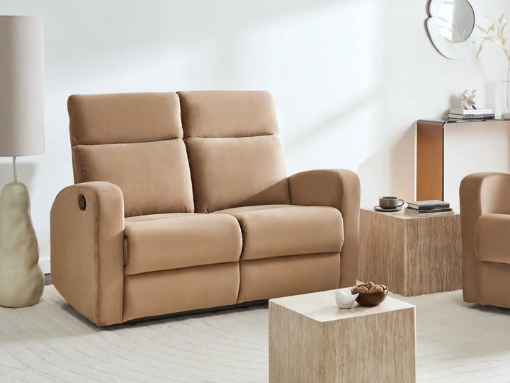 A beige upholstered reclining loveseat sofa shown in a bright, modern living room setting with a matching armchair, wooden side table, and decorative lamp.