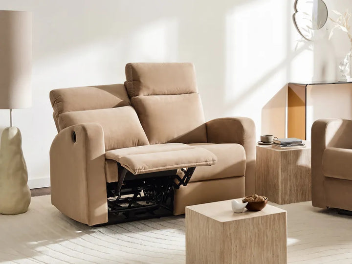 A beige upholstered recliner sofa shown in a bright, modern living room with the footrest extended. The room includes a matching armchair, wooden side tables, a floor lamp, and natural sunlight streaming across the space.