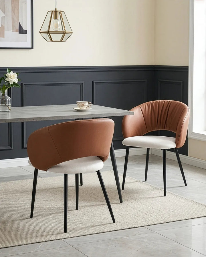Two modern dining chairs in a styled dining room setting. One chair features a warm brown leather backrest and a white upholstered seat with black legs, while the other features a matching brown leather backrest, a cream-colored seat, and black legs.