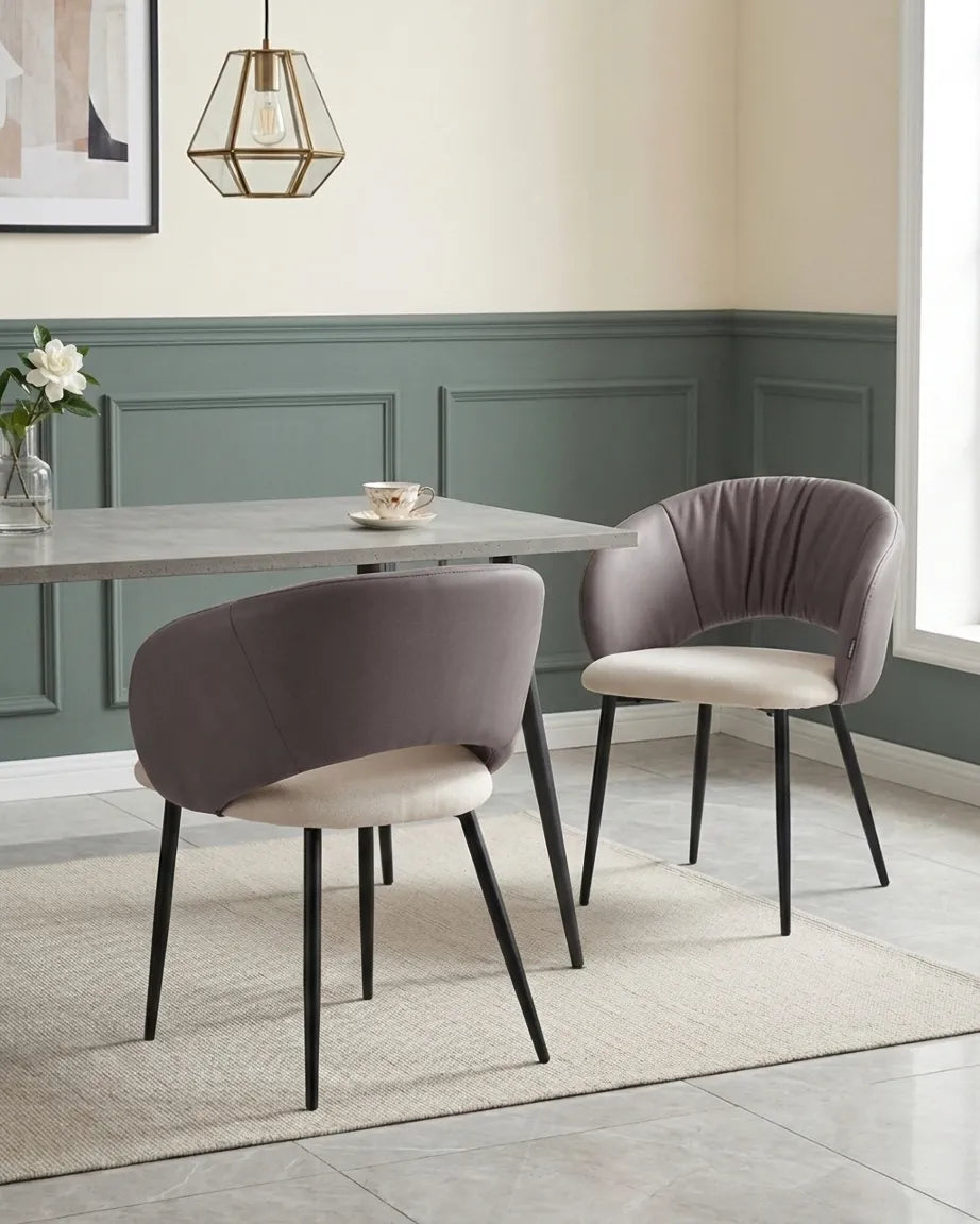 A dining room scene showing two upholstered dining chairs with curved backs, taupe-gray fabric and cream seats, black tapered legs, and a gray stone dining table in a styled interior.