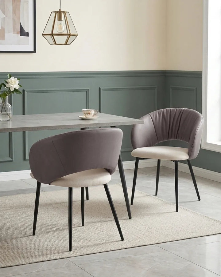 A dining room scene showing two upholstered dining chairs with curved backs, taupe-gray fabric and cream seats, black tapered legs, and a gray stone dining table in a styled interior.