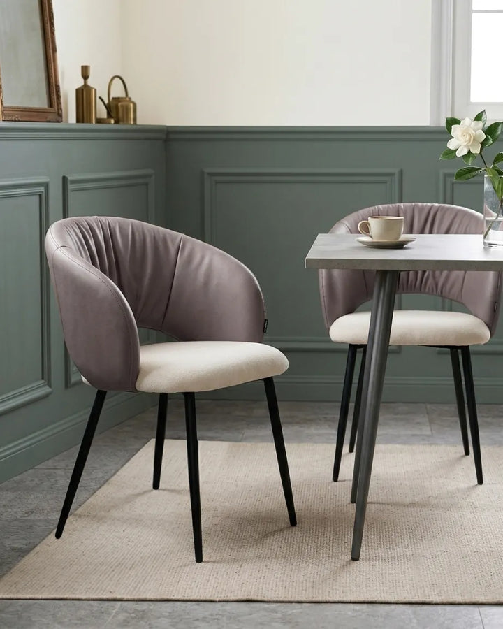 A modern dining room scene featuring two upholstered accent chairs with dusty mauve velvet backs and cream seat cushions, paired with a light wood table on a beige rug.