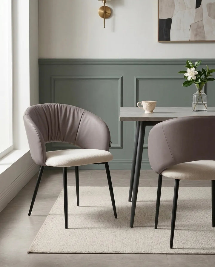 A modern dining room scene featuring two upholstered accent chairs with curved backs, one in taupe and one in light beige, paired with a gray table and a neutral rug against a green paneled wall.