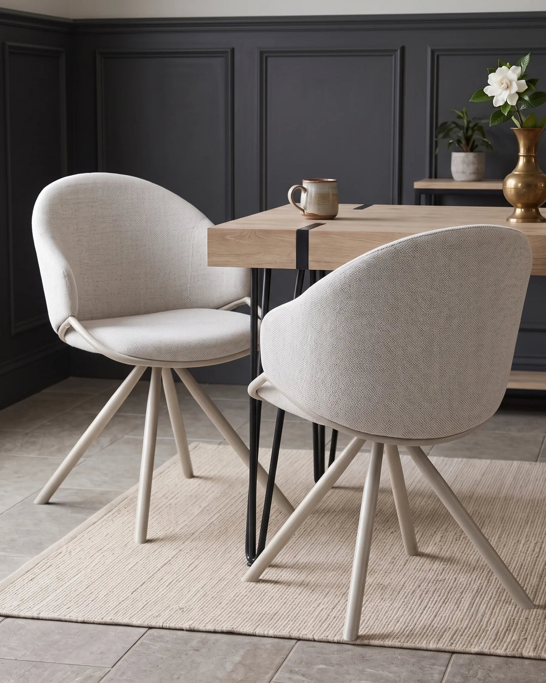 Two modern upholstered dining chairs with light beige fabric and tapered cream legs, arranged around a light wood table in a dark paneled room.