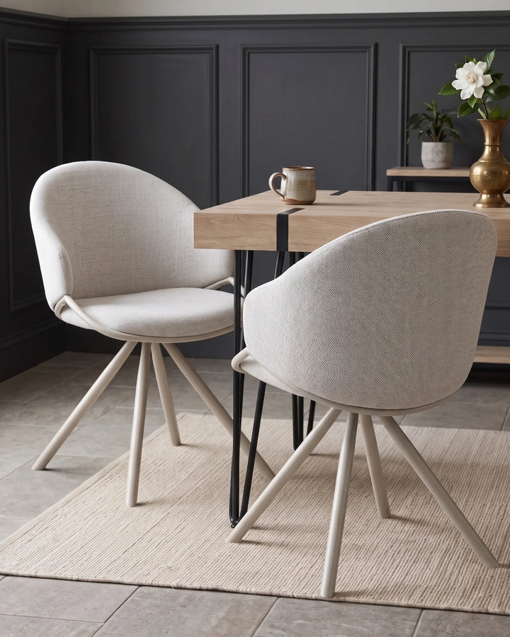 Two modern upholstered dining chairs with light beige fabric and tapered cream legs, arranged around a light wood table in a dark paneled room.