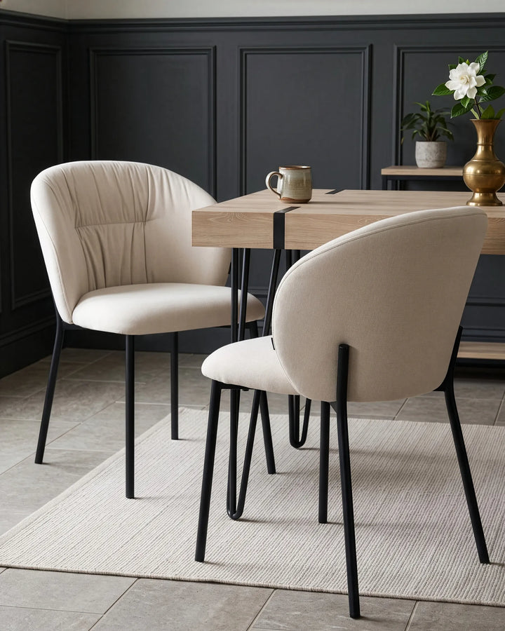 Two upholstered dining chairs with cream fabric and black metal legs, arranged around a light wood table in a modern dining room with dark paneled walls.
