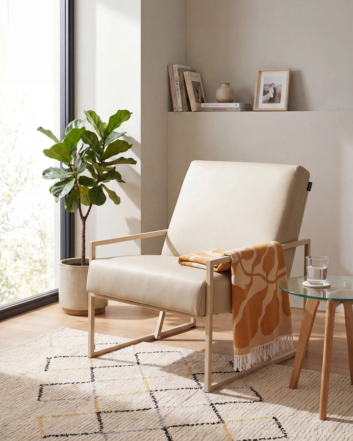 A modern cream upholstered armchair with a light wood frame sits in a bright, neutral-toned living room. A patterned orange throw blanket is draped over the arm, next to a small glass-top side table and a potted fiddle-leaf fig plant.