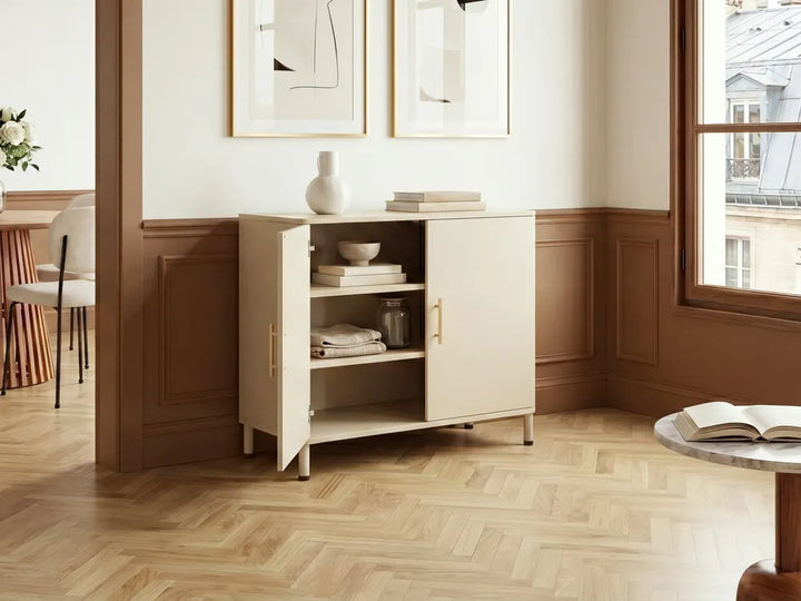 A cream-colored wooden sideboard cabinet with one open door, styled with books and a vase in a warm, modern room with wood paneling and herringbone floors.
