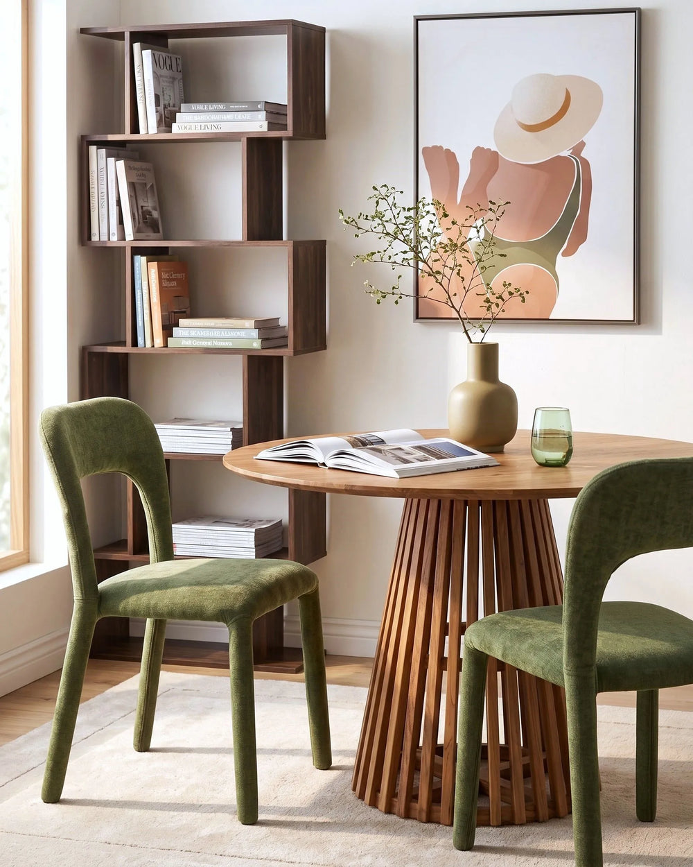A styled dining room corner featuring a round wooden slatted table, two green upholstered chairs, a tall dark wood bookshelf, and a framed art print on the wall.