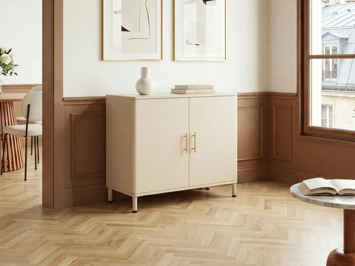 A cream-colored two-door cabinet with gold handles, styled in a warm, modern room with wood-paneled walls and a herringbone wood floor.