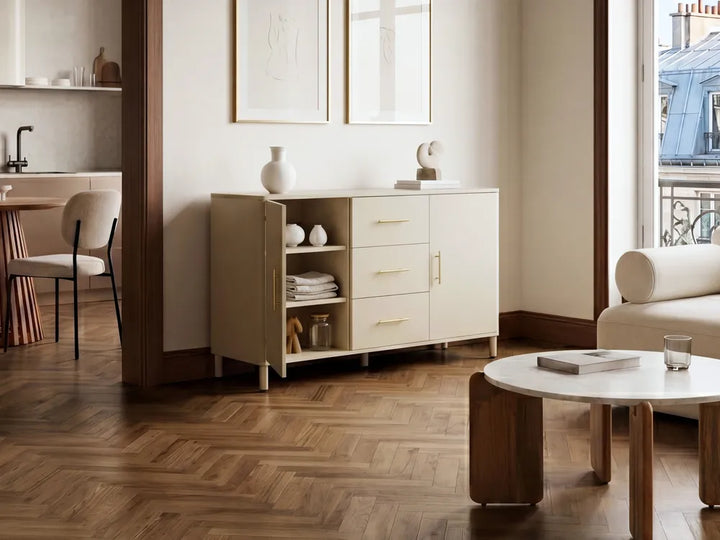 A cream-colored wooden sideboard cabinet with gold handles, styled in a modern, warm-toned living room with herringbone wood floors.