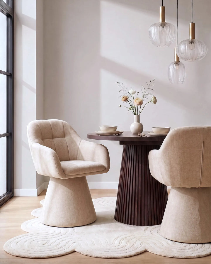A modern dining room scene featuring two upholstered dining chairs in a warm beige fabric, arranged around a dark wood round table on a sculptural cloud-shaped cream rug. A matching chair is partially visible on the right.