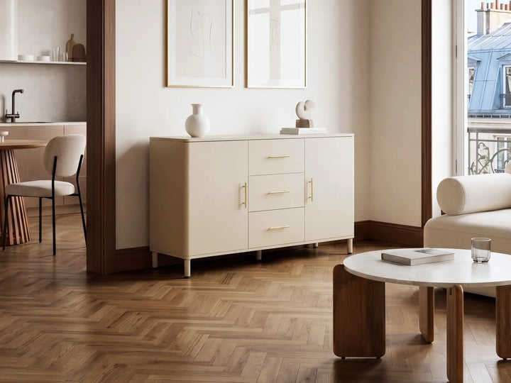 A cream-colored sideboard cabinet with three drawers and two doors, accented with slim gold handles, positioned in a modern living room with a herringbone wood floor.