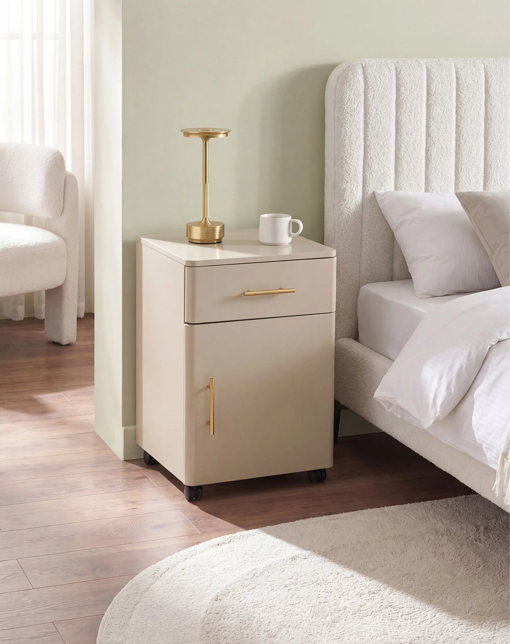 A modern beige nightstand with gold handles stands beside a bed in a softly lit bedroom. A small gold table lamp and a white mug sit on top of the nightstand.