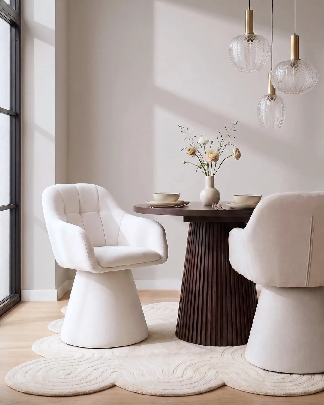 A modern dining room scene featuring two upholstered accent chairs, a round dark wood table with a vase of flowers, pendant lights, and a cloud-shaped cream rug in a bright interior.
