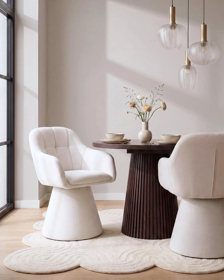 A modern dining room scene featuring two upholstered accent chairs, a round dark wood table with a vase of flowers, pendant lights, and a cloud-shaped cream rug in a bright interior.