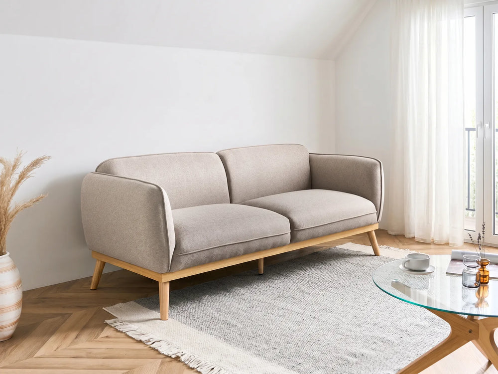 A modern two-seater sofa in light beige fabric with a natural wood frame and tapered legs, styled in a bright, minimalist living room next to a glass coffee table and a white rug.