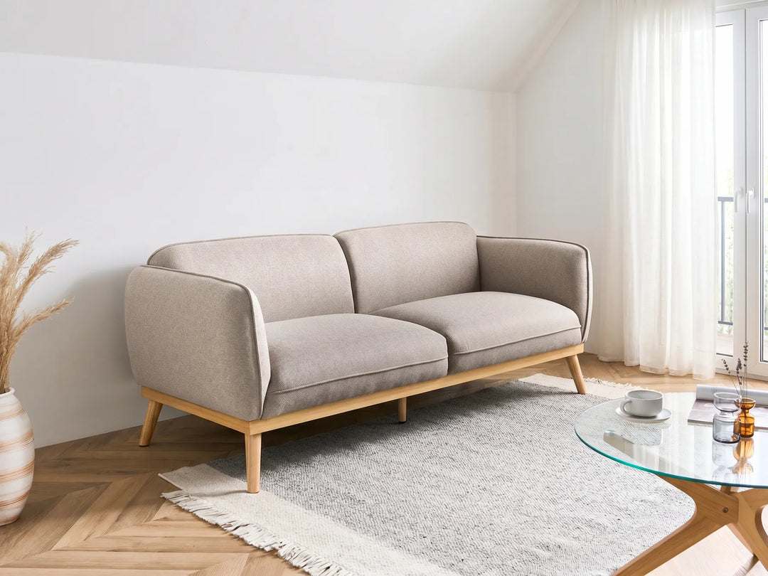 A modern two-seater sofa in light beige fabric with a natural wood frame and tapered legs, styled in a bright, minimalist living room next to a glass coffee table and a white rug.