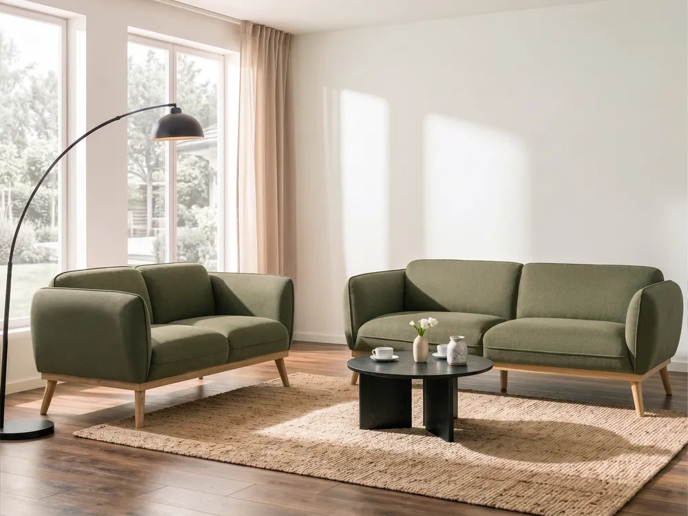 A modern living room setup featuring an olive green two-seater sofa and a matching armchair with light wood legs, arranged on a beige rug around a round black coffee table. A tall black floor lamp stands near large windows with beige curtains.