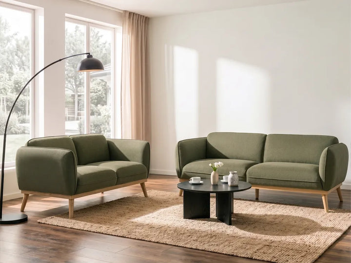 A modern living room setup featuring an olive green two-seater sofa and a matching armchair with light wood legs, arranged on a beige rug around a round black coffee table. A tall black floor lamp stands near large windows with beige curtains.
