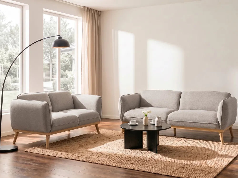 A modern living room setup featuring a light gray upholstered loveseat and a matching sofa with light wood legs, arranged on a woven beige rug around a small black coffee table. A tall black floor lamp stands near large windows with beige curtains.
