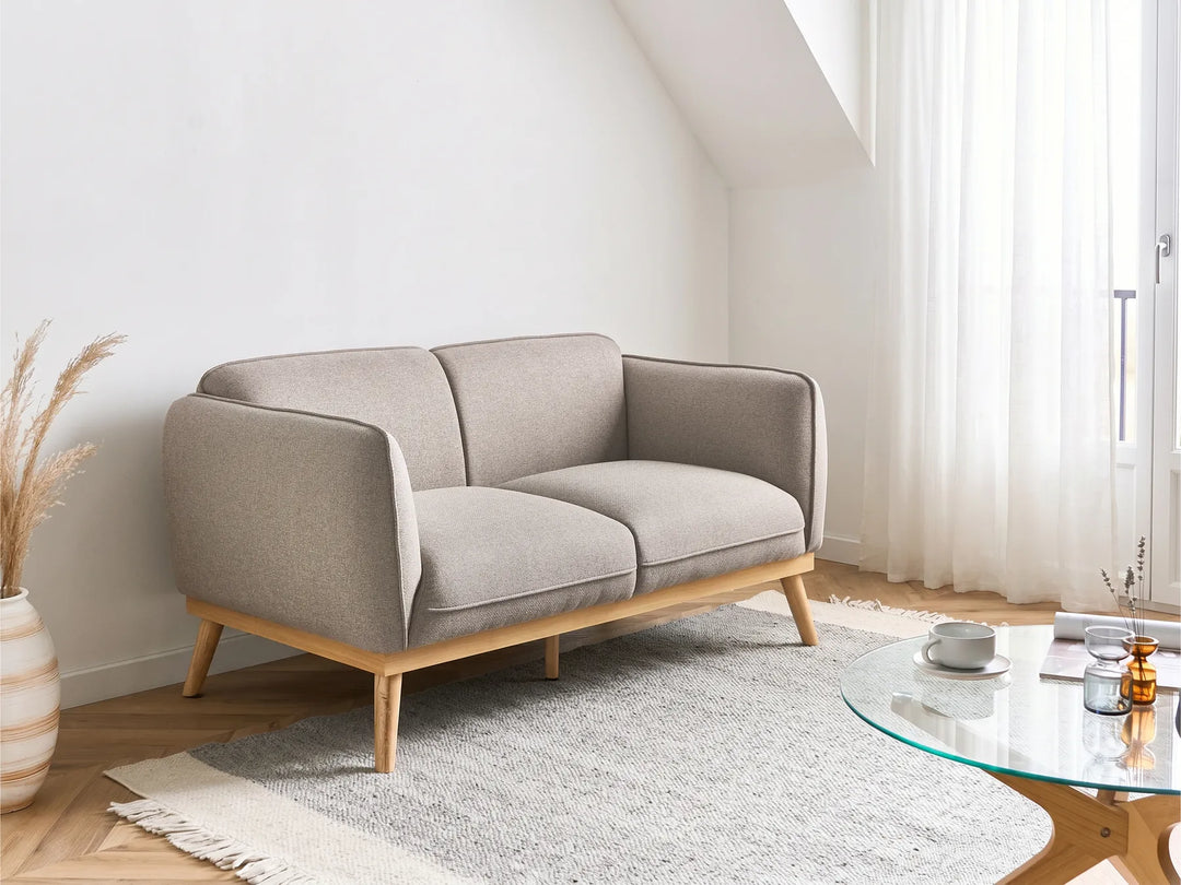 A modern two-seater sofa in light beige fabric with a natural wood frame and tapered legs, styled in a bright, minimalist living room next to a glass coffee table and a white rug.