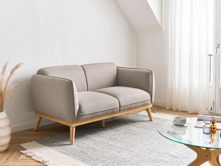 A modern two-seater sofa in light beige fabric with a natural wood frame and tapered legs, styled in a bright, minimalist living room next to a glass coffee table and a white rug.