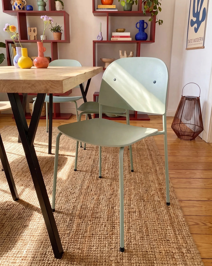 A mint green molded plastic dining chair with a minimalist design, shown in a cozy, sunlit dining room setting next to a wooden table on a woven jute rug.