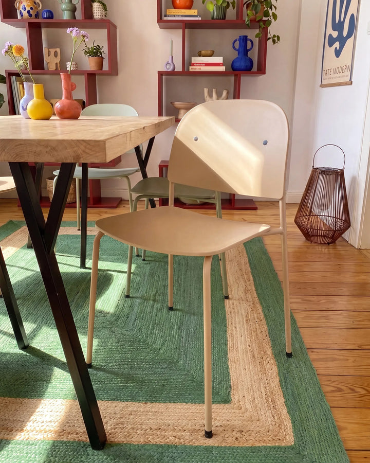A modern light beige dining chair with a curved backrest, shown in a styled dining room setting next to a wooden table on a green and beige patterned rug.