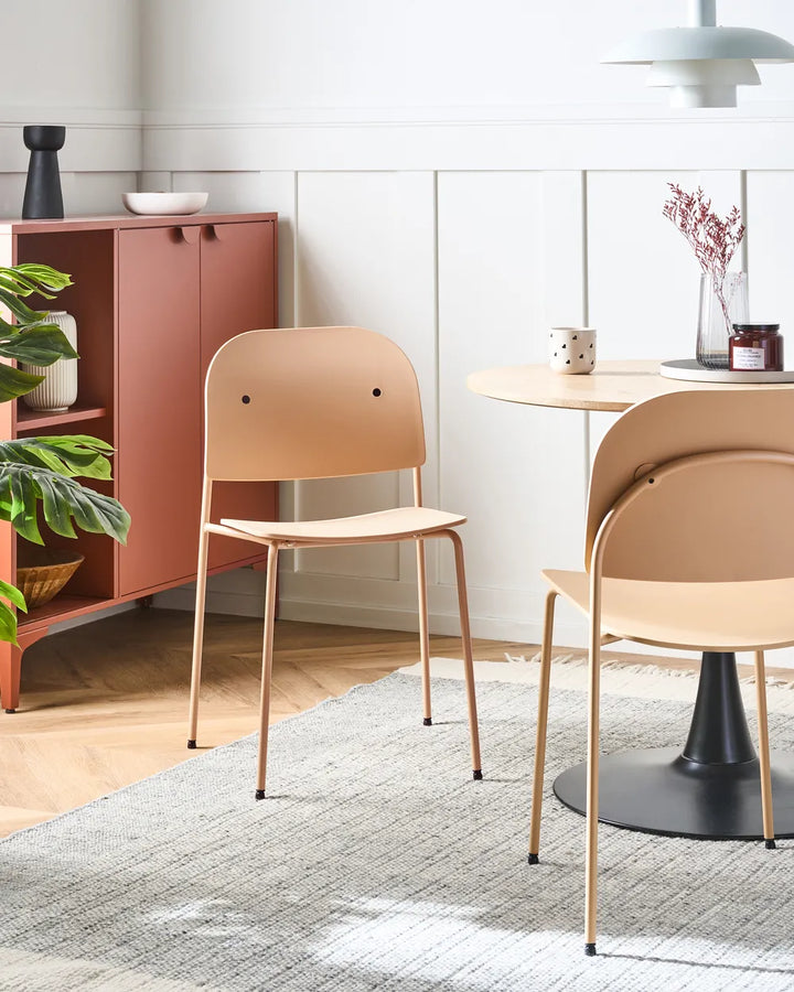 A pair of modern dining chairs in a warm beige/tan finish with slim gold-toned metal legs, shown in a styled dining room setting next to a round table and a wooden cabinet.