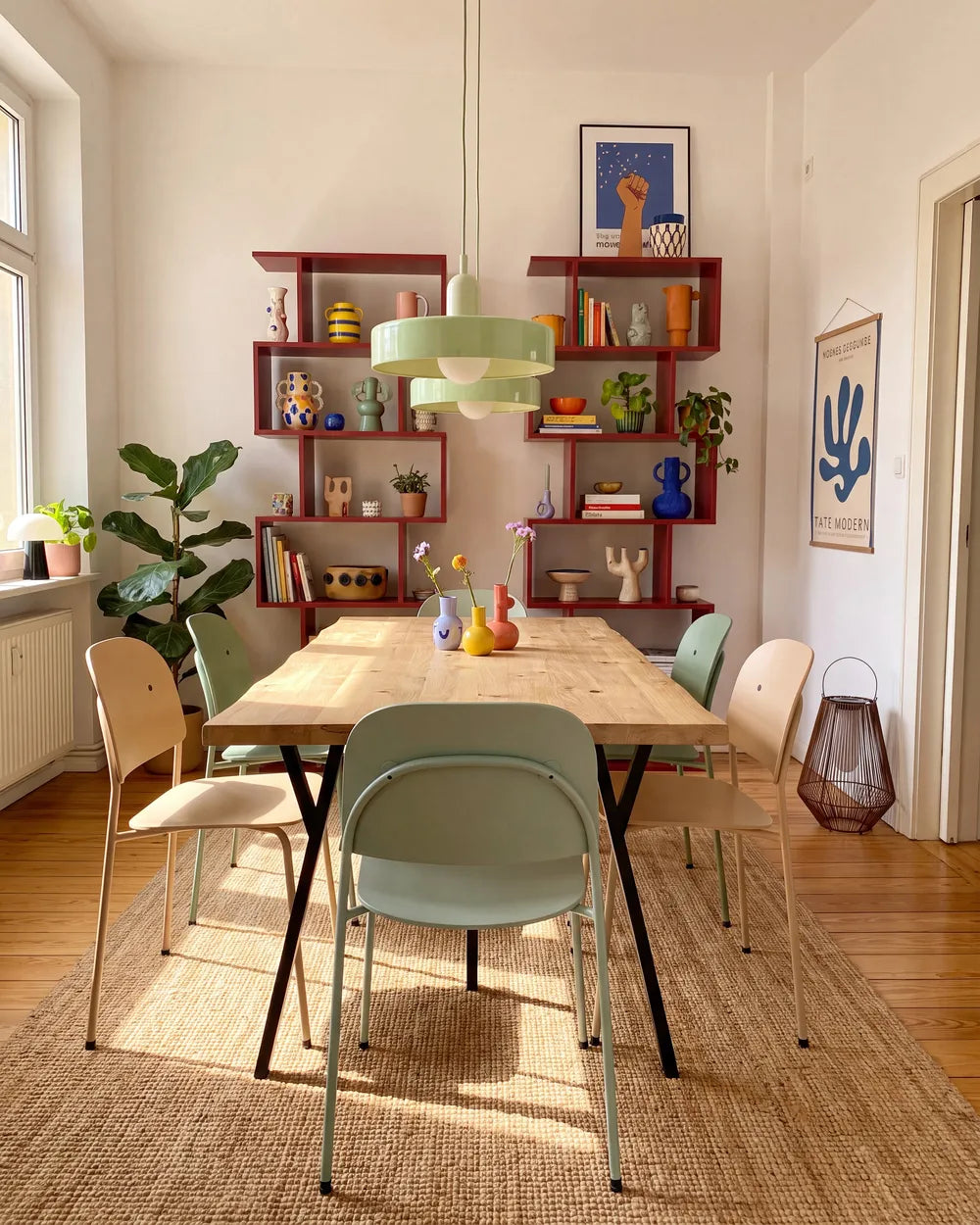 A styled dining room scene featuring a light wood dining table surrounded by pastel green, beige, and cream chairs, set on a woven jute rug. In the background, red floating shelves are filled with decorative items, and a light green pendant light hangs over the table.