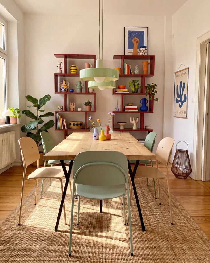 A styled dining room scene featuring a light wood dining table surrounded by pastel green, beige, and cream chairs, set on a woven jute rug. In the background, red floating shelves are filled with decorative items, and a light green pendant light hangs over the table.