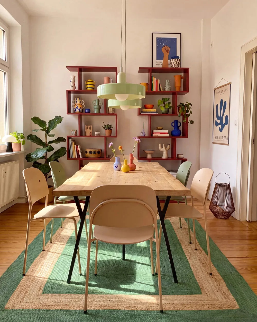 A styled dining room featuring a light wood dining table surrounded by beige and green chairs, set on a green and beige patterned rug. In the background, a large red floating bookshelf is filled with books, decorative items, and plants, under a green pendant light.