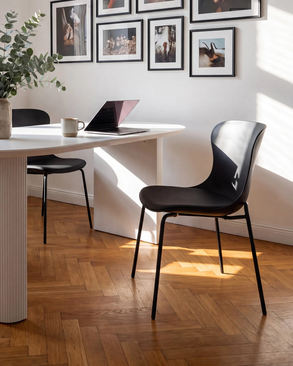 A modern black molded-acrylic dining chair with thin black metal legs, shown in a bright, sunlit room with a white table, wood herringbone floor, and framed wall art.