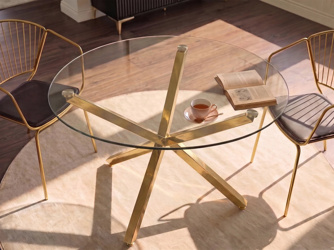 A modern round glass-top dining table with a gold-tone starburst metal base, shown in a sunlit room with two matching gold chairs and a cup on the tabletop.