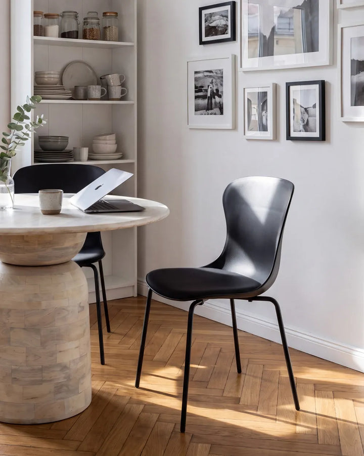 A modern black molded-acrylic dining chair with thin black metal legs, shown in a bright, sunlit dining room next to a round stone pedestal table and a white shelving unit.
