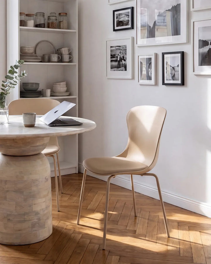 A modern beige molded plastic dining chair with slim metal legs, shown in a bright, sunlit dining room next to a round stone pedestal table.
